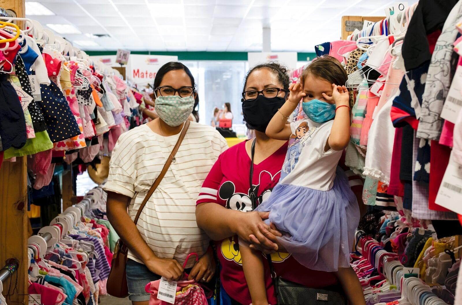 Two masked moms—one holding a child with a mask, the other pregnant—shop for their families at the local JBF sale.